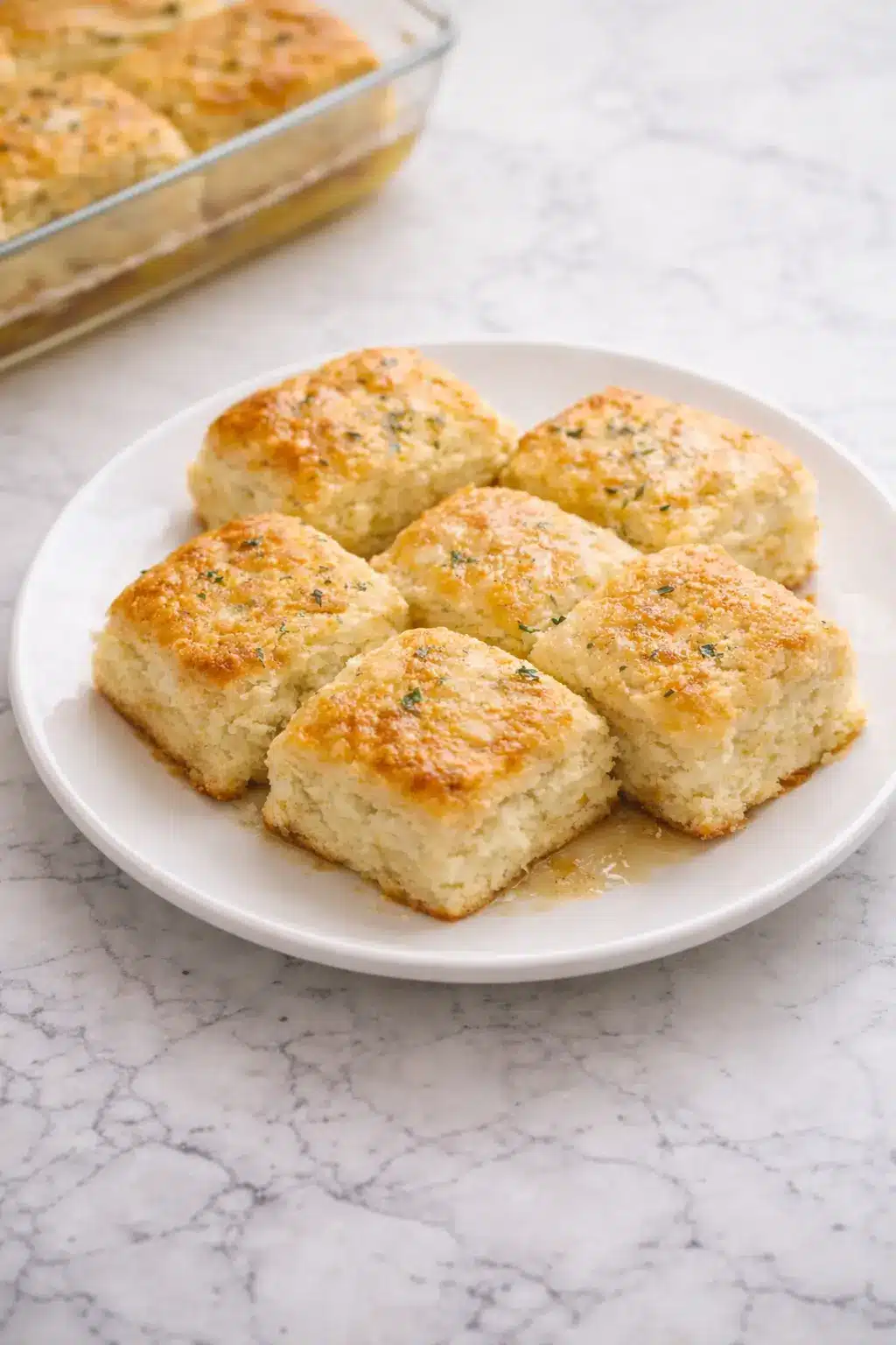 Vertical shot of golden butter swim biscuits on white plate with baking dish and parsley on marble surface.