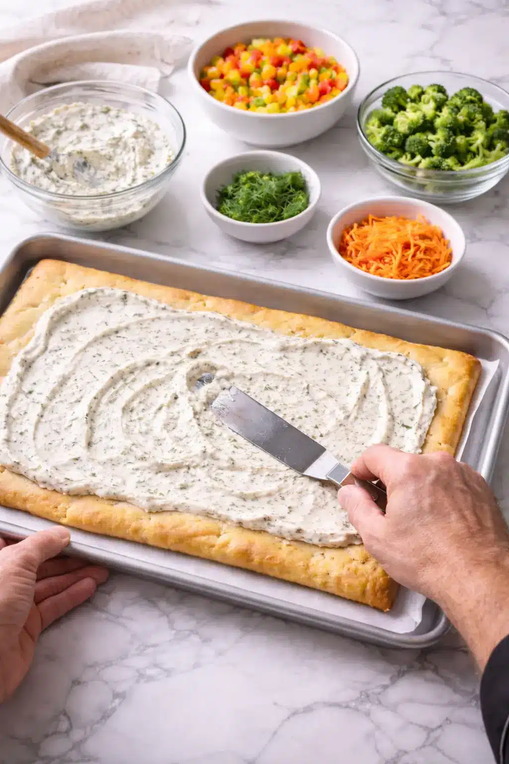 Hands spreading creamy topping on Crescent Roll Veggie Pizza crust during preparation