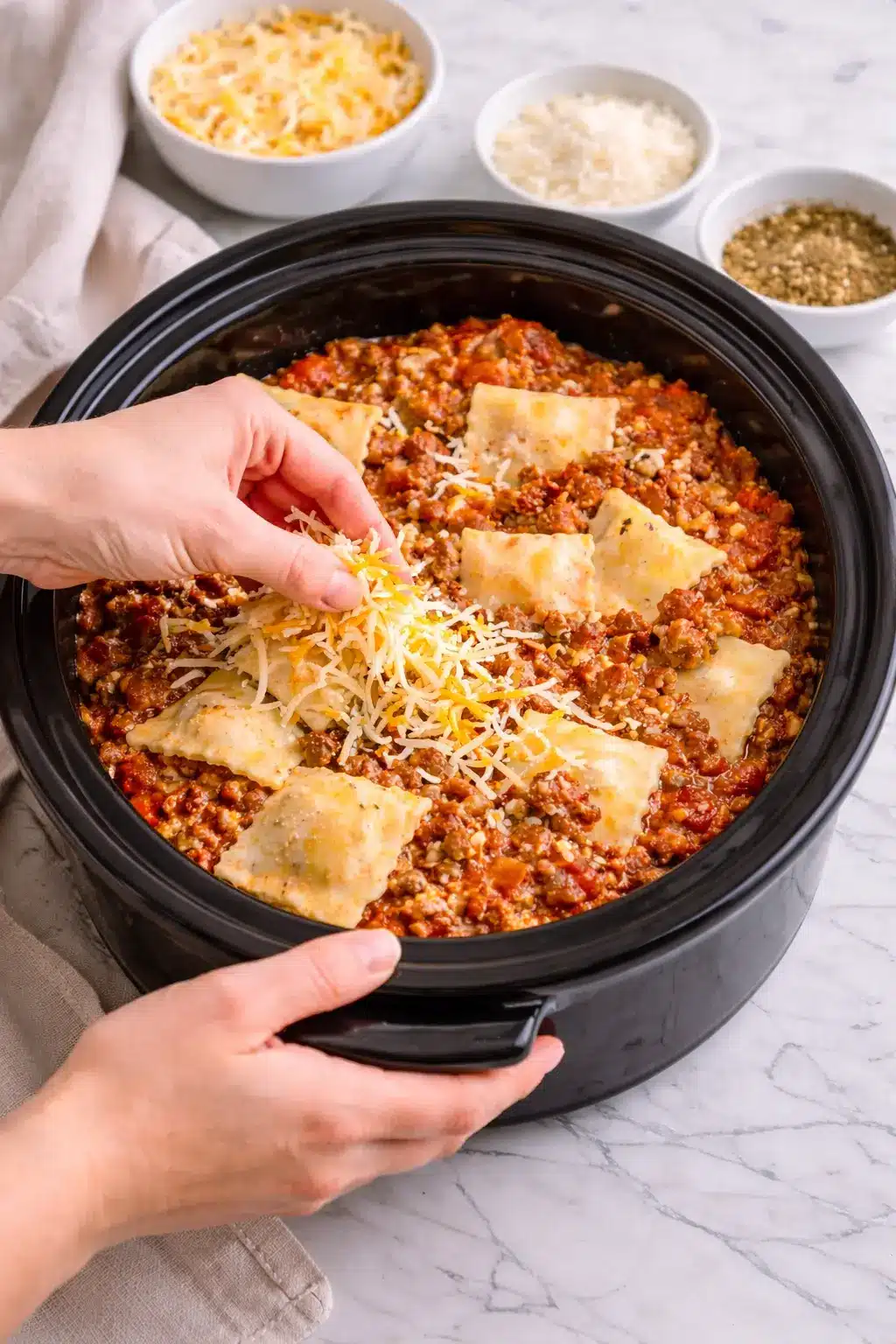 Layering Crockpot Ravioli Lasagna in a slow cooker with ravioli, meat sauce, and shredded cheese