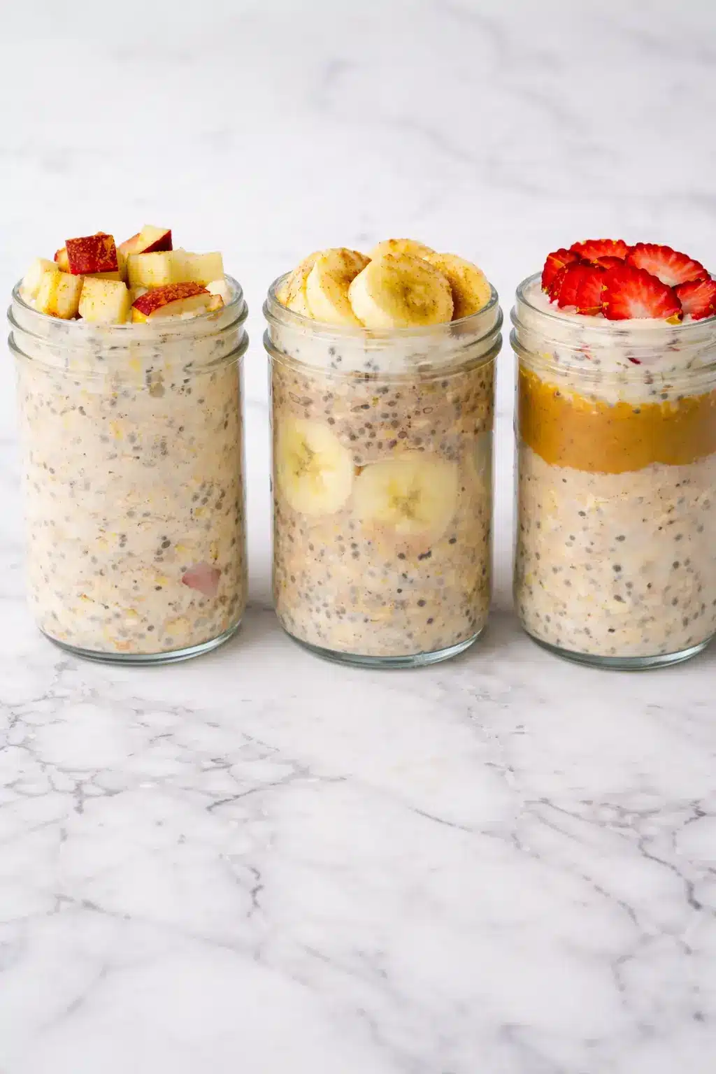High-Protein Overnight Oats in three glass jars topped with apple, banana, and strawberry slices on a white marble counter