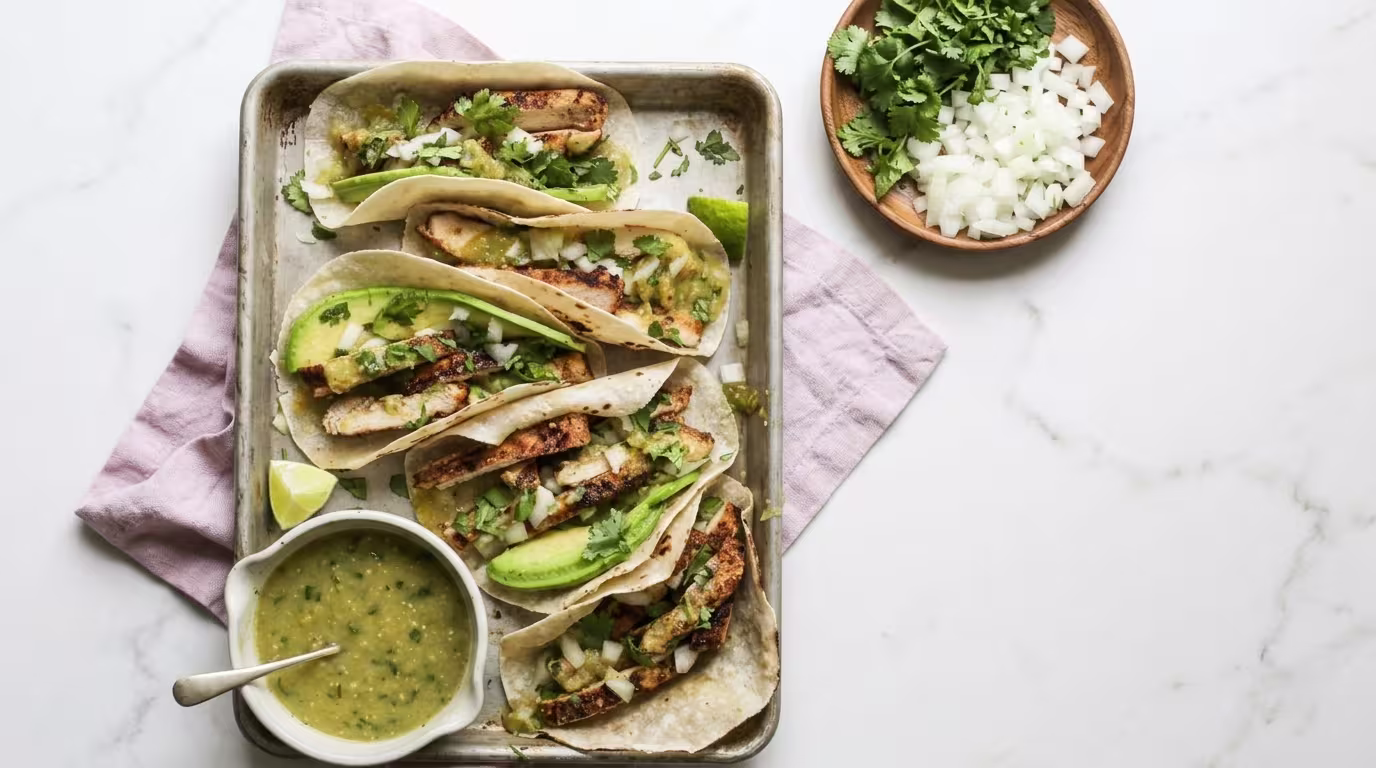A baking sheet of Tomatillo Chicken Tacos with Salsa Verde, grilled chicken strips, sliced avocado, diced onions, and fresh cilantro served on a white marble countertop with a bowl of homemade salsa verde.