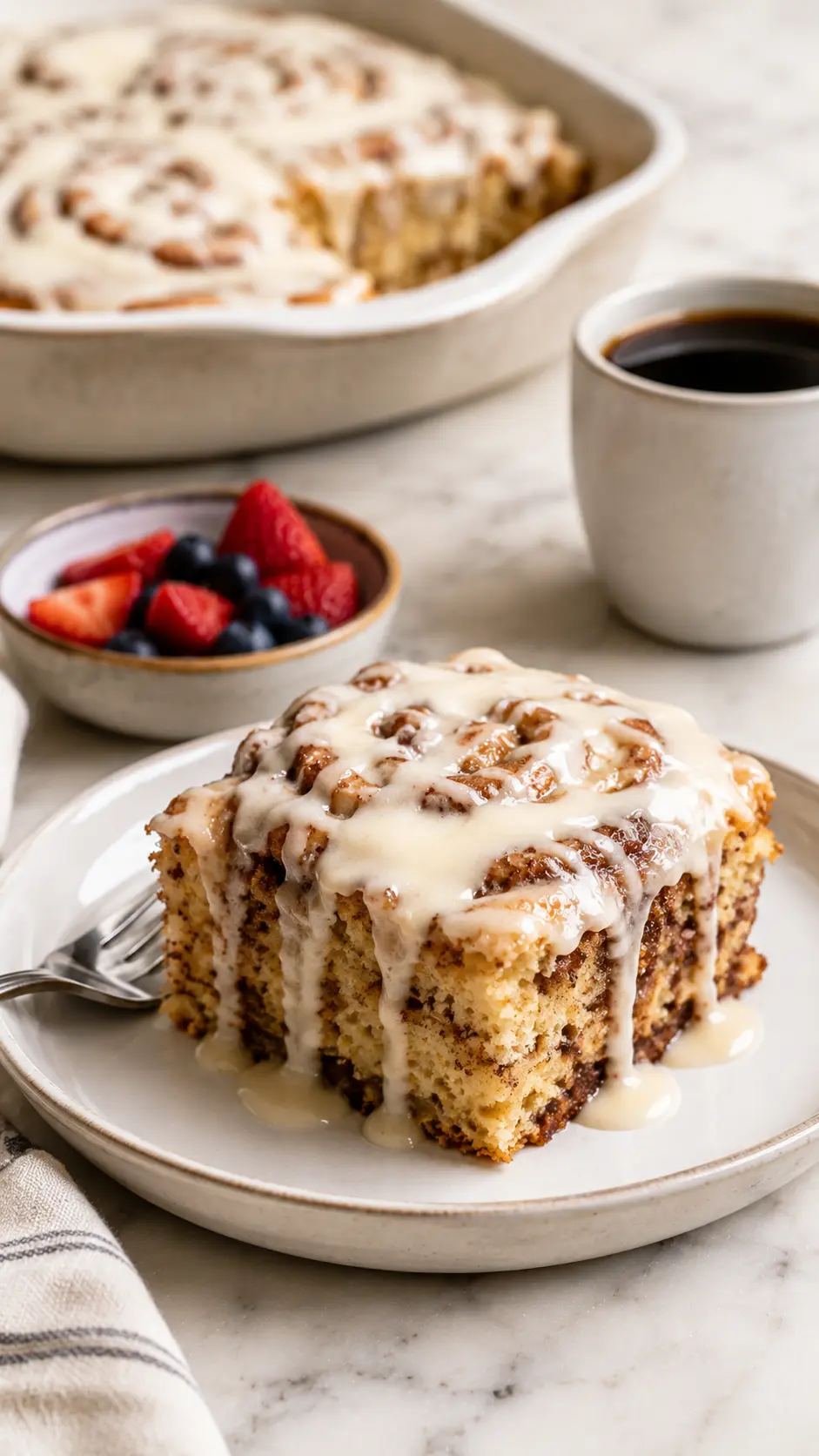 Cinnabon Cinnamon Roll Cake Cinnabon Cinnamon Roll Cake served on a plate with cream cheese icing, fresh berries, and a cup of coffee on a white marble table