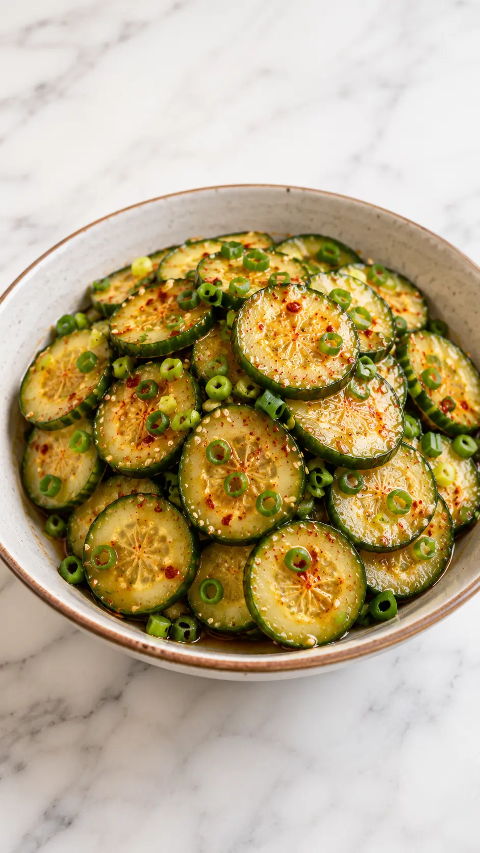 Vertical 9:16 photo of Korean Cucumber Salad with sliced cucumbers, sesame seeds, green onions, and Korean chili flakes in a ceramic bowl on white marble